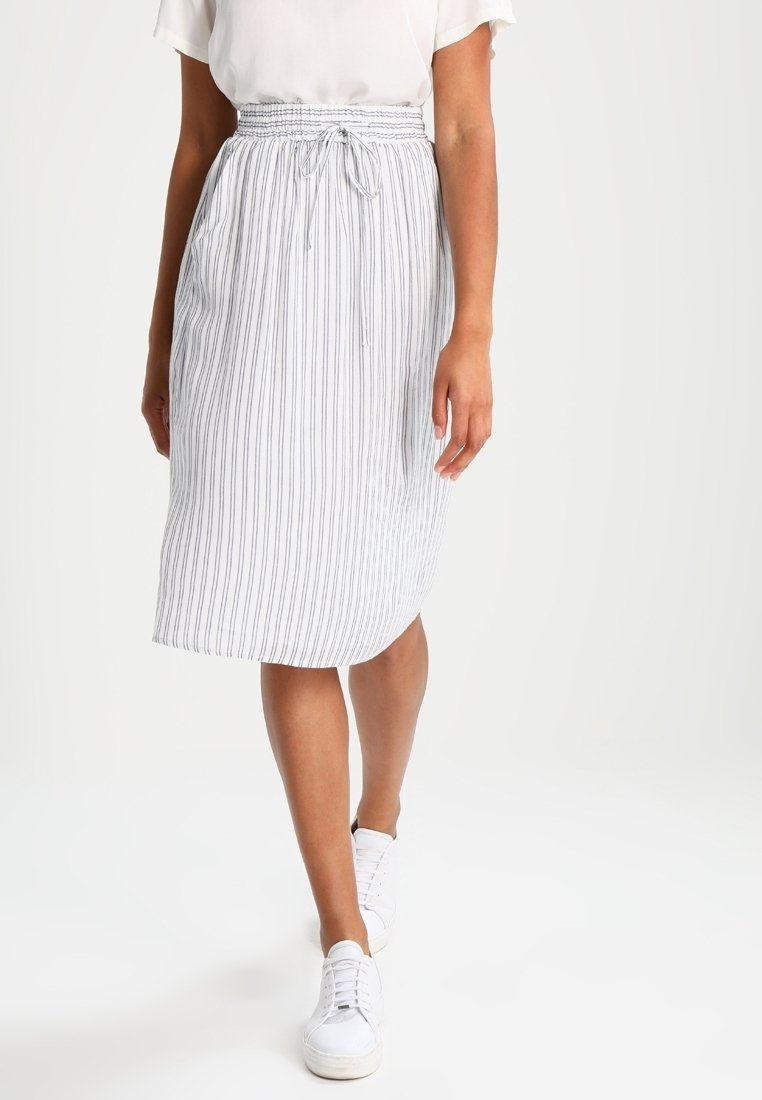 Femme portant une jupe midi à rayures verticales blanches et grises avec une taille à cordon, un haut blanc et des baskets blanches, marchant vers l'avant.
