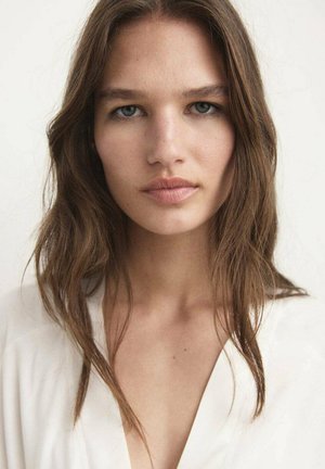 Porträt einer Person mit langen, welligen braunen Haaren, glatter Haut und dezentem Make-up. Der Hintergrund ist rein weiß. Die Kleidung besteht aus einem weichen weißen Stoff.