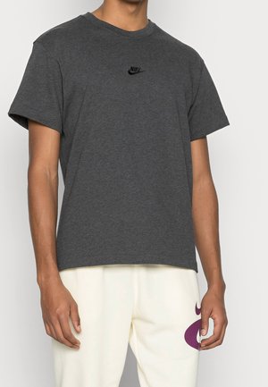Mand iført en mørkegrå Nike T-shirt og hvide bukser med et lilla design på venstre lår, stående mod en ensfarvet baggrund.