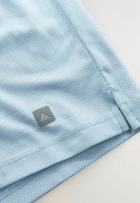 Tissu bleu clair respirant avec de petites perforations, présentant un ourlet cousu et un petit patch triangulaire gris avec logo près du bord.