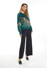 Groene trui met metallic pailletten in diagonale strepen, gecombineerd met zwarte wijde broek en zwarte hoge hakken met gouden accenten.