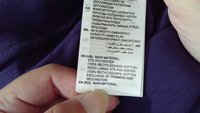 Hand holding a clothing label on purple fabric showing material details in multiple languages including English and Arabic.