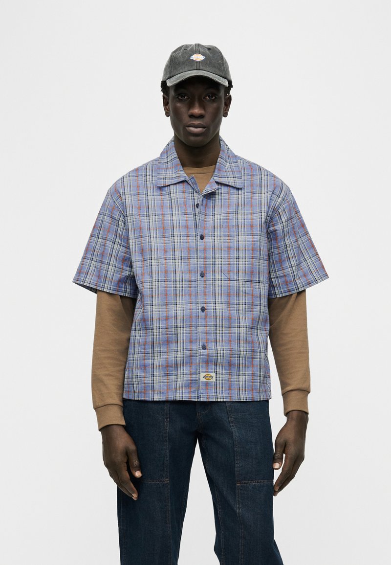 Uomo con cappellino grigio, camicia a maniche corte a quadri blu e arancioni sopra una maglia marrone a maniche lunghe, e jeans blu scuro, in piedi.