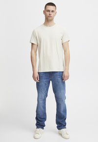 Joven de pie, vestido con una camiseta sencilla de color beige claro, jeans azules y zapatillas blancas, contra un fondo blanco.