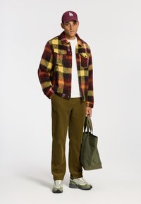 Giacca di lana a quadri rossi, gialli e neri, t-shirt bianca, pantaloni in velluto a coste olivastre, borsa a tracolla verde, sneakers grigie con dettagli bianchi, cappellino bordeaux.