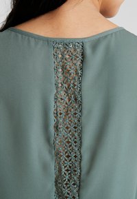 Blouse verde menta con pannello posteriore in intricate ricami di pizzo floreale, che offre un contrasto con il tessuto liscio.