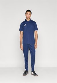 Σετ από μπλε ναυτικό πόλο και παντελόνι με λευκές λεπτομέρειες· ύφασμα που απορροφά την υγρασία· διαθέτει κουμπότρυπα και λογότυπο με τρεις ρίγες στο στήθος.