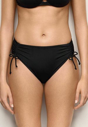 Žena nosící černé bikini kalhotky se stahováním po stranách a řasenými detaily, zobrazena od pasu po polovina stehen na jednobarevném pozadí.