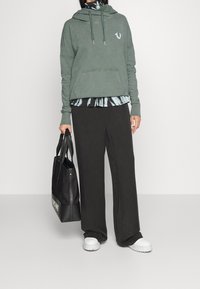 Persona che indossa un hoodie verde sopra una maglietta tie-dye, con pantaloni larghi neri e scarpe bianche, mentre tiene una grande borsa nera a tracolla.