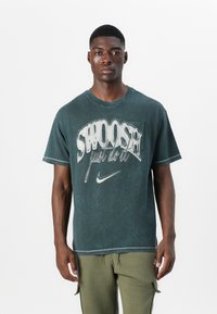 T-shirt in cotone verde foresta con un prominente design grafico bianco che presenta il testo "Swoosh" e "Just Do It"; maniche corte, vestibilità rilassata.