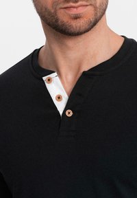 Černé tričko Henley s bílým límečkem, obsahující tři béžové knoflíky a měkkou bavlněnou strukturu. Kolo výstřih s viditelným švem.