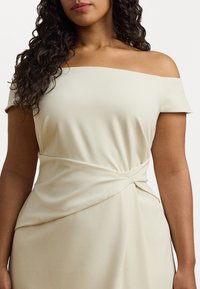 Beige off-shoulder mekko, joka on valmistettu sileästä kankaasta. Mekossa on kierretty yksityiskohta vyötäröllä ja lyhyet hihat, jotka luovat istuvan siluetin.