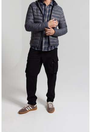 Veste matelassée grise avec capuche, portée sur une chemise à carreaux bleue et blanche, associée à un pantalon cargo noir et des baskets marron avec des rayures blanches.