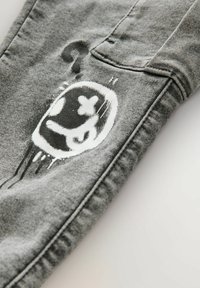 Gray denim pants featuring a distressed design with a white graphic print of a face-shaped pattern on the thigh area.