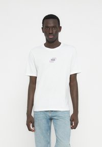 Valkoinen lyhythihainen puuvillainen T-paita, jossa on pieni suorakaiteenmuotoinen violetti- ja musta logo rinnassa, yhdistetty vaaleansinisiin farkkuihin.