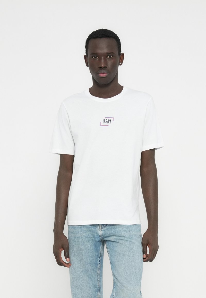 Valkoinen lyhythihainen puuvillainen T-paita, jossa on pieni suorakaiteenmuotoinen violetti- ja musta logo rinnassa, yhdistetty vaaleansinisiin farkkuihin.