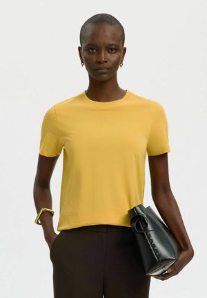 Kvinde med kort hår iført en gul kortærmet skjorte og mørke bukser, der holder en sort håndtaske og bærer guldsmykker.