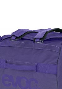 Borsa da viaggio viola con tessuto strutturato, doppie maniglie superiori e logo brandizzato. Le zip completano il design complessivo.