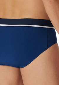 Sous-vêtement brief bleu marine avec une ceinture élastique rayée blanche, texture lisse et design ajusté, montrant la vue de côté et de dos.