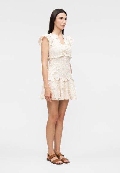 Γυναίκα που στέκεται πλευρικά φορώντας ένα αμάνικο κρεμ δαντελένιο φόρεμα με κυματιστές άκρες και καφέ επίπεδα σανδάλια σε λευκό φόντο.