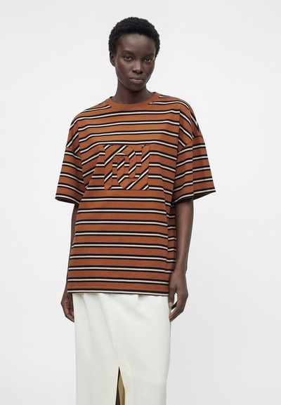 Άτομο που φοράει καφέ και μαύρη οριζόντια ριγέ oversized μπλούζα με σχέδιο στο στήθος, συνδυασμένη με λευκή φούστα με σκίσιμο μπροστά.