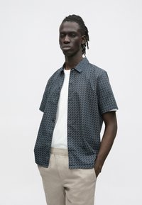 Korte mouw, marineblauw overhemd met wit zigzagpatroon, gedragen over een wit T-shirt. Gecombineerd met beige, lichtgewicht broek.