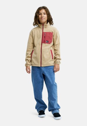 Kind met krullend haar, gekleed in een beige jas met rode accenten, blauwe spijkerbroek en zwarte sneakers, staand tegen een witte achtergrond.