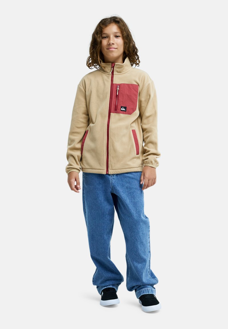Kind met krullend haar, gekleed in een beige jas met rode accenten, blauwe spijkerbroek en zwarte sneakers, staand tegen een witte achtergrond.