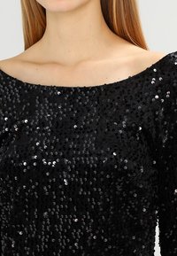 Svart paljett-topp med off-shoulder-design, med en texturerad yta täckt av glänsande, runda paljetter för ökad visuell intress.