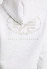 Lysegrå hoodie med en blød tekstur, der har et stort logo på ryggen og teksten "ALPHA INDUSTRIES" i en subtil nuance.