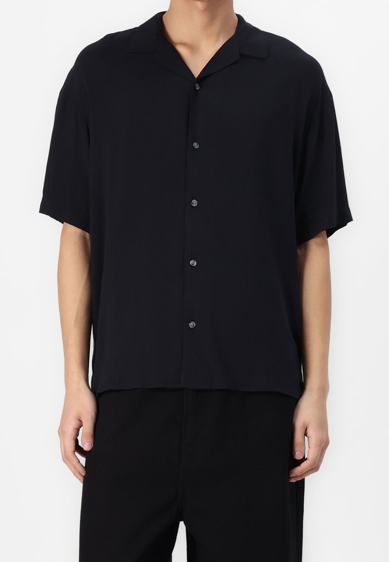 Camisa negra de manga corta en tejido ligero, con cuello tipo solapa y cierre de botones, con un corte holgado y una textura suave.