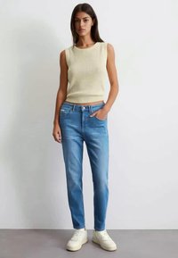 Bežinė juostelėmis megzta be rankovių palaidinė derinama su šviesiai mėlynomis tiesiomis kelnėmis. Aprangą papildo baltos sportbačiai.