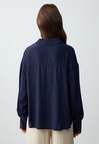 Marineblauw langemouwen shirt met een kraag, ontspannen silhouet, gatherende mouwen en een subtiele textuur. Achteraanzicht toont eenvoudige ontwerpelementen.