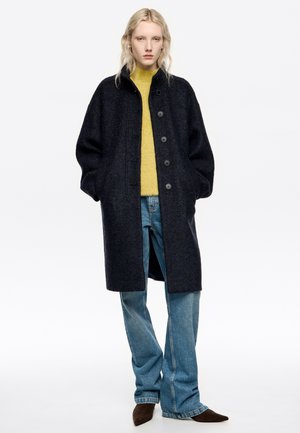 Abrigo de lana gris oscuro con un corte relajado, que presenta un cuello alto y cinco botones. Combinado con un suéter amarillo brillante y jeans de cielo azul.