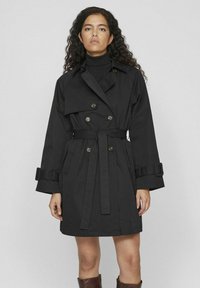 Svart trenchcoat med dubbla knappar, vida ärmar, midjebälte och två framfickor. Slät tyg med minimal detaljering.