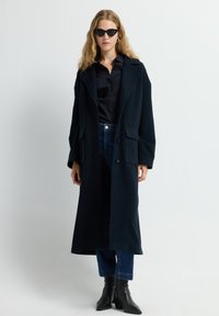 Dunkelblauer, gestreifter langer Mantel mit Revers, Seitentaschen und zwei Knöpfen, getragen über einem schwarzen Hemd und blauen Jeans, kombiniert mit schwarzen Knöchelstiefeln.