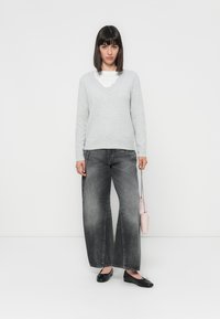 Vrouw met steil, donker haar, gekleed in een lichtgrijze trui, wijdvallende vervaagde zwarte spijkerbroek, zwarte flats en houdt een kleine roze handtas vast.