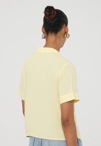 Camicia a maniche corte con colletto in tessuto giallo pallido dalla consistenza morbida. Presenta polsini piegati e un orlo leggermente accorciato.