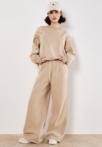 Felpa beige con ricami floreali sulle maniche, abbinata a pantaloni larghi coordinati. Tessuto testurizzato e vita con cordoncino regolabile.