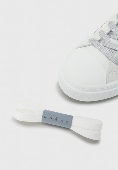 Baskets blanches avec une tige en cuir lisse, des lacets gris et une semelle en caoutchouc texturée, accompagnées de lacets blancs supplémentaires avec une étiquette grise.