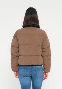 Femme aux cheveux raides et foncés, portant une veste matelassée brune et un jean bleu, de dos devant un fond blanc uni.