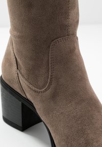 Detailní záběr na taupe semišovou kotníkovou botu s propracovaným švem a černým blokovým podpatkem na bílém pozadí.