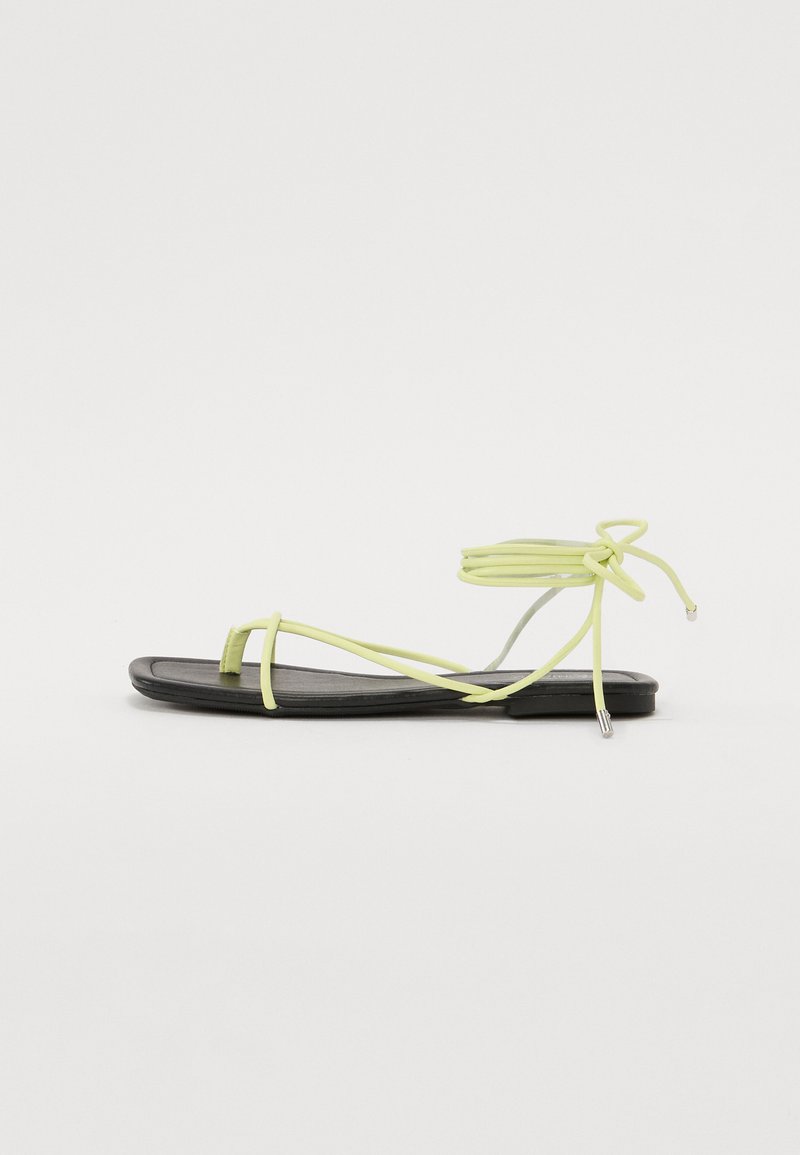 Keltaiset hihnasandaalit mustalla litteällä pohjalla. Ominaisuudet ohuet hihnat ja nilkkasideet, valmistettu sileästä materiaalista, jossa on minimalistinen muotoilu.