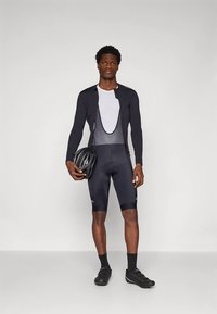 Ποδηλατική ενδυμασία που περιλαμβάνει εφαρμοστό μαύρο μπλουζάκι με μακρύ μανίκι και σορτς, με λευκό δίχτυ στη ράχη. Το μοντέλο κρατάει ένα μαύρο κράνος.