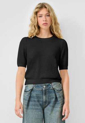 Blonde vrouw die een zwart, gestructureerd, kortemouwentruitje draagt en blauwe high-waisted jeans met een unieke dubbele knoopdetail.