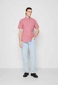 Chemise à manches courtes à carreaux rouges et blancs avec une patte de boutonnage, associée à un jean bleu clair et des chaussures noires. Texture de tissu lisse.