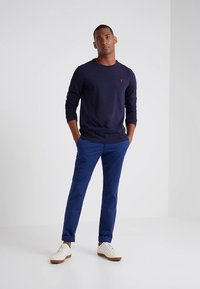 Tengerészkék hosszú ujjú ing, világoskék chino nadrág és fehér tornacipő. Az ing mellrészén kis piros logó látható. Egyszerű, sima textúrák mindenütt.