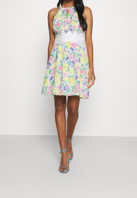 Robe à col licou florale avec une taille en dentelle blanche ajustée et une jupe évasée, présentant des couleurs pastel en rose, jaune et violet. Bretelles réglables.