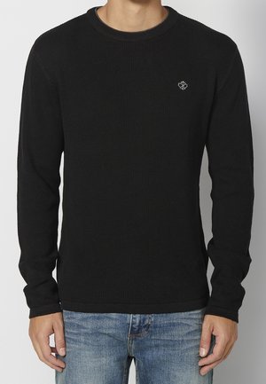 Uomo che indossa un maglione a maniche lunghe nero a coste con un piccolo emblema a foglia bianca sul petto e jeans azzurri chiari.