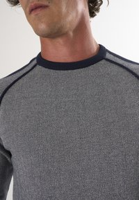 Textured grijze sweater met een marineblauwe geribde ronde halslijn en schouderaccenten. Close-up weergave toont het gedetailleerde breiwerk.